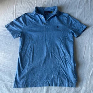 Blå pikétröja från Polo Ralph Lauren - Säljer en klassisk blå pikétröja från Polo Ralph Lauren i modellen Custom Slim Fit. Tröjan har korta ärmar, krage och knappar framtill samt den ikoniska broderade loggan på bröstet. Perfekt för en clean och stilren look.