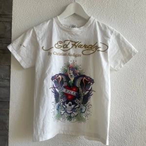 Vit Ed Hardy t-shirt med tryck - Vit t-shirt från Ed Hardy med ikoniskt färgstarkt tryck framtill med ormar, dödskalle och hjärta. Guldigt logotryck både fram och bak. Klassisk passform och rund hals. Materialet är mjuk bomull, baby T-shirt, perfekt för en edgy streetstil.   #90s #stockholm #skate#trending #y2k #baggy #triftfinds  #cottagecoreaesthetic #pintereststyle #itgirl #swag #unique #90s #00s #2000s #y2kfashion #aesthetic #streetstyle #streetwear #lowwaist #lowrise #cool #skater #baggyfit 