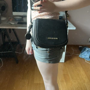 Svart axelväska från Michael Kors - Snygg svart axelväska från Michael Kors med justerbar rem och guldfärgade detaljer. Väskan är i skinn och har ett stilrent, klassiskt utseende med logga framtill. Perfekt storlek för det viktigaste och passar till många olika outfits.