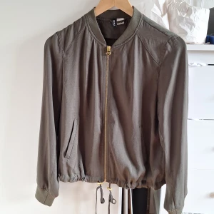 Olivgrön kofta från H&M Divided - Snygg olivgrön kofta från H&M Divided med guldfärgad dragkedja, ribbade muddar och dragsko nertill. Koftan har två fickor och är i ett lätt material som passar perfekt till vår och höst. Enkel och stilren design som funkar till många outfits.