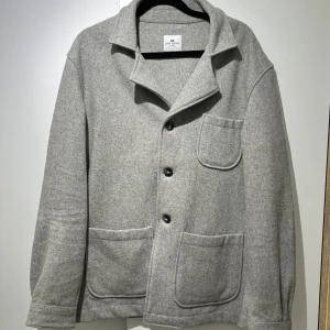 Grå overshirt/kavaj - John Henric - Stilren grå overshirt från John Henric med tre stora fickor framtill och klassisk krage. Jackan har knäppning med svarta knappar och är tillverkad i mjukt ullmaterial. Perfekt för lager-på-lager och en clean look.