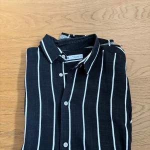Svartvit randig skjorta från Zara - Svart skjorta med vita vertikala ränder från Zara. Skjortan har korta ärmar, klassisk krage och knäppning framtill med vita knappar. Tillverkad i ett lätt och luftigt linnematerial, perfekt för varma dagar. Snygg och stilren design som passar till många outfits.