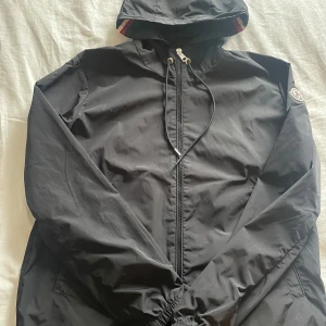 Svart vindjacka från Moncler med huva - Svart vindjacka från Moncler med klassisk logga på ärmen. Jackan har huva med snörning och dragkedja framtill. Lätt och smidig modell i syntetmaterial, perfekt för blåsiga dagar. Snygga detaljer med Monclers signaturfärger på insidan av huvan.