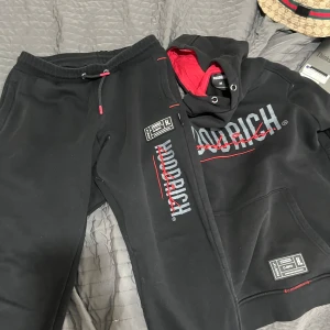 Svart HOODRICH hoodie med röd detalj - Svart hoodie från HOODRICH i storlek M med stor logga framtill i grått och rött. Hoodien har röd insida i huvan, snörning och en stor magficka. Snygga detaljer med etikett på fickan och cool streetstil.