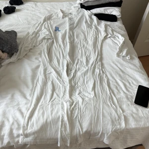 Vit henley-skjorta från Martinson - Säljer en vit henley-skjorta från Martinson med lång ärm och knappar framtill. Skjortan har en enkel, rak passform och ett blått tryckt JL-märke på bröstet. Tillverkad i mjuk bomull, perfekt för en clean och avslappnad look.