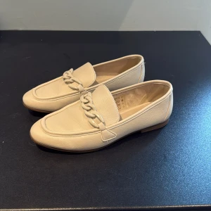 Beige loafers med kedjedetalj från Din Sko - Stilrena beige loafers i skinn med chunky kedjedetalj framtill. Skorna har platt sula, rund tå och en klassisk siluett som passar till allt. Perfekta för dig som gillar en clean och modern look med en twist.