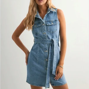 Ärmlös jeansklänning med bälte - Snygg ärmlös jeansklänning i ljusblå denim med knappar framtill, krage och två bröstfickor. Klänningen har ett matchande knytbälte i midjan som ger en schysst siluett. Perfekt för dig som gillar en avslappnad men trendig look. aldrig använd och den är slutsåld på Nelly hemsidan 