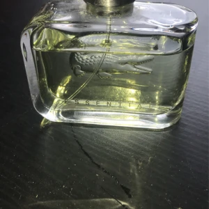 Lacoste Essential parfym 75ml - Fräsch parfym från Lacoste, Essential, i en stilren rektangulär glasflaska med silverfärgad metallkork. Flaskan har den klassiska krokodilloggan ingraverad och innehåller en ljusgul vätska. Perfekt för dig som gillar sportiga och fräscha dofter.