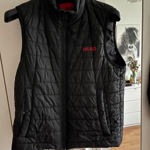 Svart quiltad dunväst från Hugo - Snygg svart dunväst från Hugo med diskret quiltat mönster och röd logga på bröstet. Västen har dragkedja framtill och två fickor med knappstängning. Perfekt att slänga över en hoodie för en clean look.