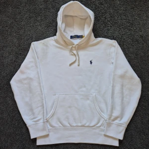 Vit hoodie från Polo Ralph Lauren - Vit hoodie från Polo Ralph Lauren med klassisk broderad logga i mörkblått på bröstet. Tröjan har huva med snörning, känguruficka och ribbade muddar. Tillverkad i mjuk bomull, perfekt för en chill och stilren look. Den har bara blivit använd 2 gånger innan det tyvärr blev en fläck på högra axeln, jag har inte lyckas få bort den men går säker bort med lite ansträngning.