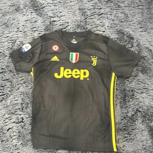 Juventus svart bortatröja Adidas - Juventus bortatröja i svart med gula detaljer och Jeep-logga på bröstet. Adidas-märke och klubbmärke framtill, samt Serie A-märke på ärmen. Tröjan är kortärmad och tillverkad i ett lätt, ventilerande material.