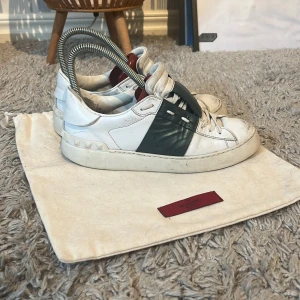 Valentino Garavani Open sneakers vit/grön - Snygga Valentino Garavani Open sneakers i vitt skinn med grön bred rem över mitten och coola nitar på hälen. Klassisk rund tå, vita snören och röda detaljer på insidan. Perfekta för dig som gillar lyxiga och trendiga sneakers med unik design. Skriv om du har några frågor!😊