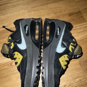 Nike Air Max 90 svart/gul sneakers - Nike Air Max 90 sneakers i svart och grått med gula detaljer och reflekterande swoosh. Ovandel i textil och mocka, mesh på plösen och tåpartiet. Klassisk Air Max-sula med synlig luftbubbla och robust yttersula. Perfekta för dig som gillar streetstyle.