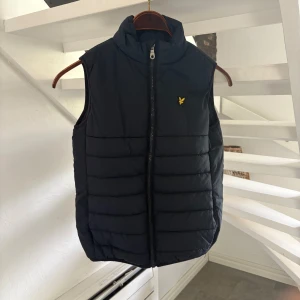 Marinblå dunväst från Lyle & Scott Junior - Snygg dunväst från Lyle & Scott Junior med gul logga på bröstet. Västen har hög krage, quiltad design och dragkedja framtill och fickor. Bra skick! 