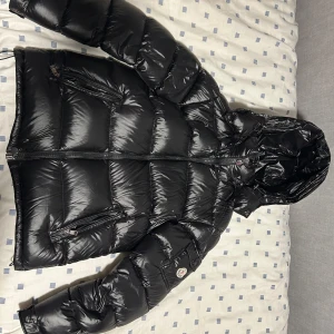 Svart Moncler pufferjacka med huva - Säljer en svart, glansig pufferjacka från Moncler med huva och dragkedja. Jackan har flera fickor med dragkedja och klassisk Moncler-logga på ärmen. Perfekt för kalla dagar när du vill hålla stilen och värmen. Fodrad insida med coolt serietryck. 