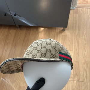Gucci keps  - Snygg beige keps från Gucci med klassiskt GG-monogram över hela kepsen. Har grönt och rött band på sidorna och justerbar rem baktill i brunt läder. Perfekt accessoar för en trendig look.