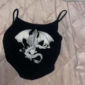 Svart korsettopp med draketryck Shein - Cool svart korsettopp från Shein med smala axelband och ett stort silverfärgat drakmotiv framtill. Toppen har en croppad passform och är tillverkad i mjukt syntetmaterial. Perfekt för dig som vill sticka ut med edgy detaljer.