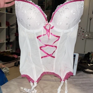 Vit och rosa korsett från Dikaru - Supersöt vit korsett från Dikaru med rosa detaljer och snörning framtill. Kuporna har broderade blommor och spetskant, och nederkanten är dekorerad med rosa volang. Justerbara axelband och hyskknäppning bak. Perfekt för dig som vill ha en feminin och lekfull look.