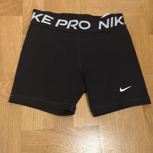 Nike Pro svarta träningstights-shorts - Svarta Nike Pro shorts med tight passform och bred resår i midjan med Nike-logga. Perfekta för träning, tillverkade i stretchigt syntetmaterial som andas. Klassisk vit swoosh på benet och stilren design som sitter på plats under intensiva pass.