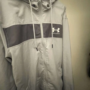 Grå vindjacka från Under Armour - Snygg grå vindjacka från Under Armour med svart panel över bröstet och vit logga. Jackan har huva med dragsko, meshfoder och hel dragkedja framtill. Perfekt för dig som gillar sportig stil och vill ha något lätt och funktionellt.