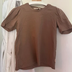 Brun kortärmad topp från Pernilla Wahlgren - Säljer en brun kortärmad topp från Pernilla Wahlgren i storlek XS. Toppen har puffiga ärmar och rund halsringning.
