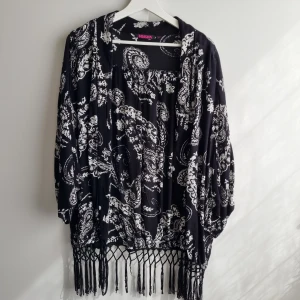 Svart kimono kofta Indiska - Svart och vit kofta från Indiska med paisley mönster och långa ärmar. Snygga fransdetaljer nertill och en loose passform. Perfekt att slänga på sig över en t-shirt för en chill och bohemisk vibe. 100% viskos 