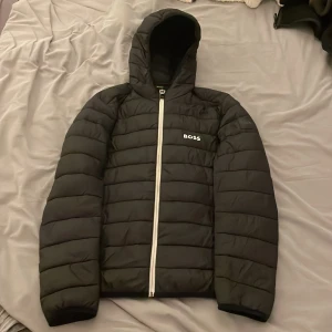 Svart pufferjacka från BOSS med huva - Svart pufferjacka från BOSS med vit dragkedja och broderad logga på bröstet. Jackan har huva, quiltad design och långa ärmar. Två skador på vänster arm en som är lite större och en en liten bredvid BOSS märket, se bild nedan. Priset är inte hugget i sten så kom med bud! Passar perfekt för blåsiga dagar men även lite kallare och sena sommarkvällar! Storleken är M, jag är 1,84 cm lång och den sitter perfekt på mig! En liten gåva medföljer🎁😆