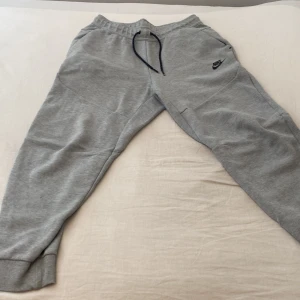 Grå mjukisbyxor från Nike - Grå mjukisbyxor från Nike med svart logga på låret. Byxorna har resår och snörning i midjan samt svarta dragkedjor på fickorna. Tillverkade i mjukt bomullsmaterial och har en relaxed passform med muddar vid bensluten.