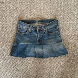 Jeanskjol med volang från Nudie Jeans Co - Säljer en snygg jeanskjol från Nudie Jeans Co i klassisk blå denim. Kjolen har volang nertill och femficksmodell med orangea kontrastsömmar. Perfekt för dig som gillar en cool och avslappnad stil.