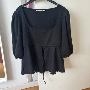 Svart blus med knyt i midjan från lager 157 - Svart blus med fyrkantig ringning och trekvartsärmar. Blusen har ett knytband i midjan och lätt puffade ärmar. Perfekt att styla med jeans eller kjol för en trendig look.