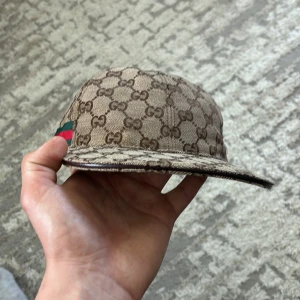 Gucci keps med GG-mönster och läderdetalj - Snygg keps från Gucci i beige och brunt med klassiskt GG-monogram över hela kepsen. Justerbar rem i mörkbrunt läder baktill och ikoniska gröna/röda ränder på sidan. Tillverkad i bomullscanvas med exklusiv känsla. Perfekt för dig som vill sticka ut med lyxig streetstyle.
