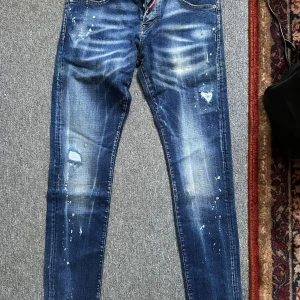 Dsquared2 blå skinny jeans med slitningar - Snygga blå jeans från Dsquared2 med skinny passform. Jeansen har coola slitningar, färgstänk och en tvättad look. Klassisk femficksmodell med knappar i gylfen och Dsquared2-logga på röd etikett vid fickan. Perfekta för en trendig streetstyle. Helt ny! 