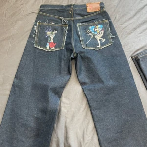 RMC Red Monkey Company Martin Ksohoh Lot1001 Men’s Jeans Diamond Ladies - Unika blå jeans med bred passform och broderade motiv på bakfickorna. Ena fickan har en broderad figur med röd sarong och lila eld, den andra en samuraj med svärd i färgglad kimono. Tuffa slitningar runt fickorna för extra edge.