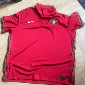 Portugal fotbollströja från Nike - Röd Portugal fotbollströja från Nike med broderad landslagslogga på bröstet och guldfärgad Nike-logga. Tröjan har korta ärmar och gröna samt röda detaljer längs sidorna. Klassisk krage och tillverkad i lätt funktionsmaterial.