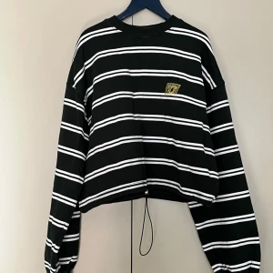 Randig sweatshirt från Junkyard - Säljer en mörkgrön croppad tröja med vita horisontella ränder och en liten gul broderad detalj på bröstet. Tröjan har långa ärmar och en avslappnad passform. Perfekt för en cool och avslappnad stil.