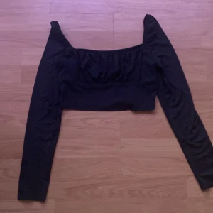 Svart croppad topp med lång ärm från SHEIN - Säljer en svart croppad topp från SHEIN med långa ärmar och rynkade detaljer vid bysten. Toppen har en fyrkantig urringning och är tillverkad i ett stretchigt material. 
