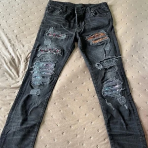 Svarta jeans med färgglada slitningar - Säljer ett par svarta jeans med färgglada tygdetaljer bakom slitningarna på framsidan. Byxorna har fem fickor, normal passform och raka ben. Perfekta för dig som vill sticka ut med en unik stil!