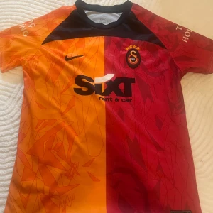 Galatasaray matchtröja Nike röd/orange - Galatasaray fotbollströja från Nike med klassisk halvorange och halvröd design, klubbmärke och sponsortryck på bröstet. Tröjan har svarta detaljer vid halsen och subtila grafiska mönster. Tillverkad i lätt och ventilerande polyester, perfekt för fotbollsplanen.
