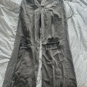 Svarta  jeans med hål på knäna - Säljer ett par högmidjade svarta jeans med stora slitna hål över båda knäna. Jeansen har en rak passform och är tillverkade i klassiskt denimtyg. Perfekta för en avslappnad och edgy stil. 