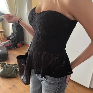 Svart spetsig bustier-topp - Säljer en svart bustier-topp med spetsdetaljer och peplum-nederdel. Toppen är axelbandslös och har en figurnära passform som framhäver siluetten. Perfekt att styla med jeans eller kjol för en trendig look.