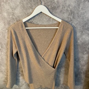 Beige ribbad omlottopp - Säljer en beige ribbad omlottopp med v-ringning och trekvartsärm. Toppen har en figurnära passform och är tillverkad i ett mjukt, stretchigt material som passar perfekt till både jeans och kjol.