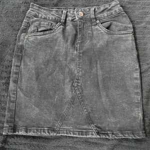 Svart jeanskjol med fickor - Säljer en svart, tvättad jeanskjol med klassiska fickor fram och bak samt knapp och dragkedja i midjan. Kjolen har en rak passform och synliga sömmar framtill som ger en cool look.