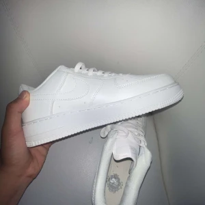 Nike Air Force 1 helvita sneakers - Klassiska Nike Air Force 1 sneakers i helvitt skinn med lågt skaft. Snygg och clean design med perforerad tå, vit sula och vita skosnören. Perfekta för dig som gillar en stilren look och vill ha ett par ikoniska sneakers i garderoben.