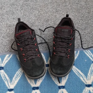 Svarta sneakers från ECCO med röda detaljer - Snygga svarta sneakers från ECCO med coola röda detaljer och svarta snören med mönster. Skorna har en robust sula och ovandel i mocka och syntet, perfekt för dig som gillar en sportig och stilren look. Låg modell med rund tå och diskret ECCO-logga på plösen.