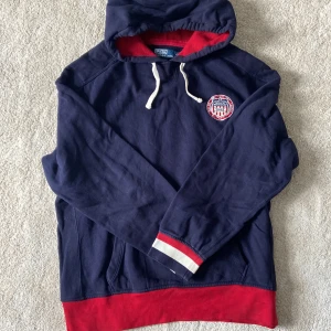 Mörkblå hoodie från Polo Ralph Lauren - Mörkblå hoodie med röd insida av huvan och nederkant, samt vit dragsko. Bröstet har ett broderat märke med texten 'United States R.L.P.C Winter Event 1934'. Perfekt för en sportig och avslappnad stil.