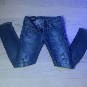 Dondup george jeans - Säljer ett par blå jeans från Dondup med slitna detaljer och råa kanter. Jeansen har klassisk femficksdesign, synliga slitningar framtill och bakfickor med kontrastsömmar. Perfekta för en avslappnad och trendig look.