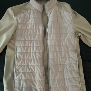 Beige quiltad jacka från ZARA - Snygg beige jacka från ZARA med quiltade paneler framtill och dragkedja. Jackan har lång ärm och en stilren, sportig look. Perfekt för lager-på-lager under kyliga dagar.