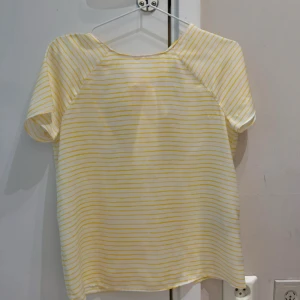 Gul- och vitrandig blus med knyt i ryggen - Säljer en söt blus med gula och vita ränder. Blusen har korta ärmar och en snygg öppen rygg med knytband upptill. Perfekt för varmare dagar och ger en fräsch look.