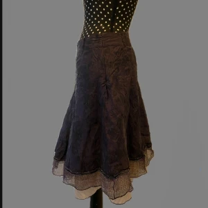 Lila medium kjol med volangkant - lätt glansigt tyg och volangkant nertill. Kjolen har ett subtilt mönster och extra lager i nederkanten med rutiga detaljer. Original pris 1200kr ärvt av min mor. Det går att ha bälte och den är rätt låg midjad. (Jag är 85-90cm) Dubbel lager så man ser inte igenom och den sitter fint på. 