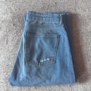 Blå jeansbyxor från Djerf Avenue - Säljer ett par klassiska blå jeans från Djerf Avenue med bakficka och liten broderad detalj med hjärta. Perfekta för dig som gillar enkel och stilren denim med en söt touch.